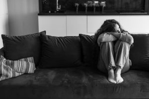 Black and white photograph of a person sitting curled up against the head of a bed, knees drawn in.