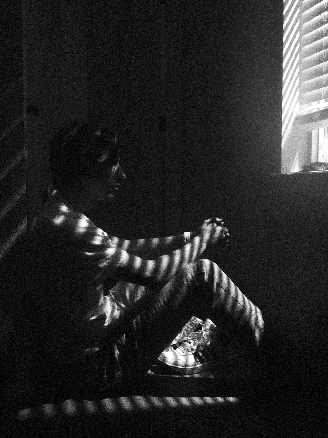 A child sitting alone on the floor beside a window with knees drawn up, striped light from a blind falling across them