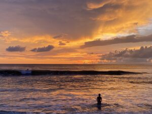 A single figure standing waist-deep in the sea, facing a wide orange sunset.