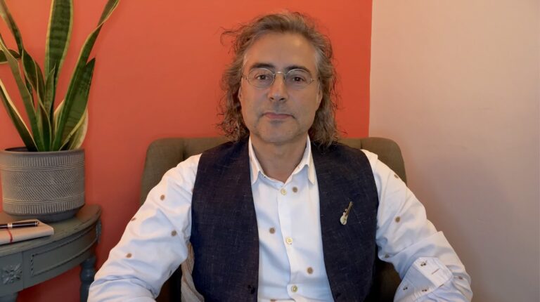 Alex Golding sitting in his consulting room, a plant to one side and a deep orange wall behind him.