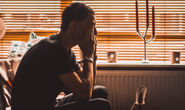 A person sitting on the floor at home, leaning back against a radiator with one hand in their hair.