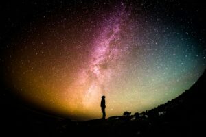A small figure standing alone beneath the Milky Way, arms slightly raised.