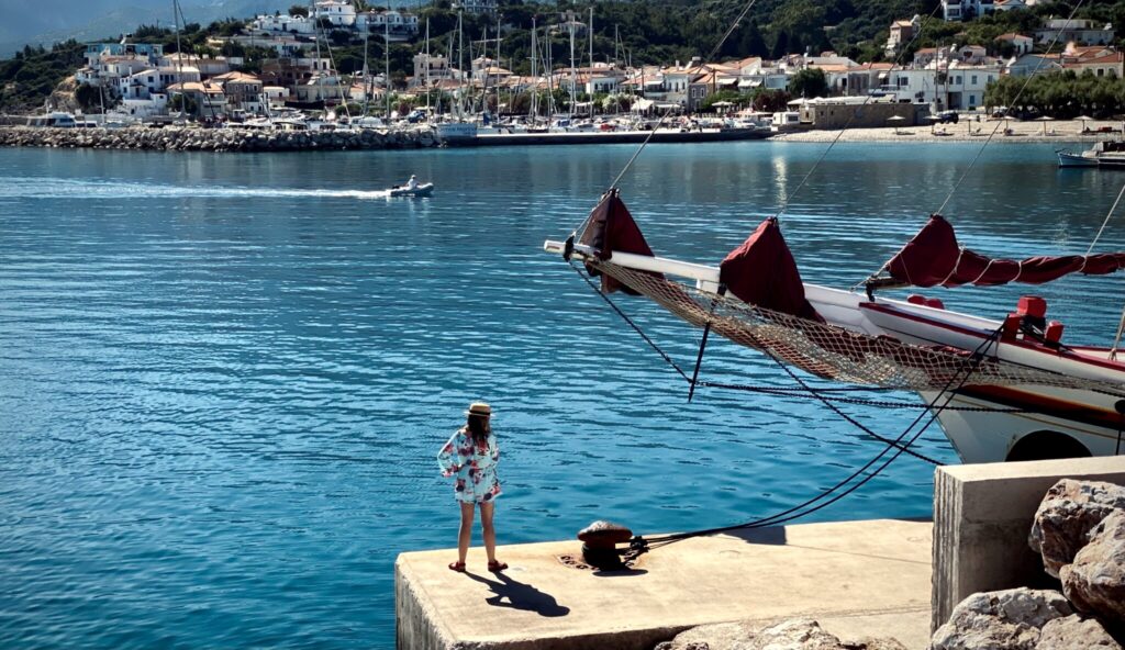 A woman standing alone at the end of a harbour wall beside a moored sailing ship, looking out across the water to the town on the far shore.