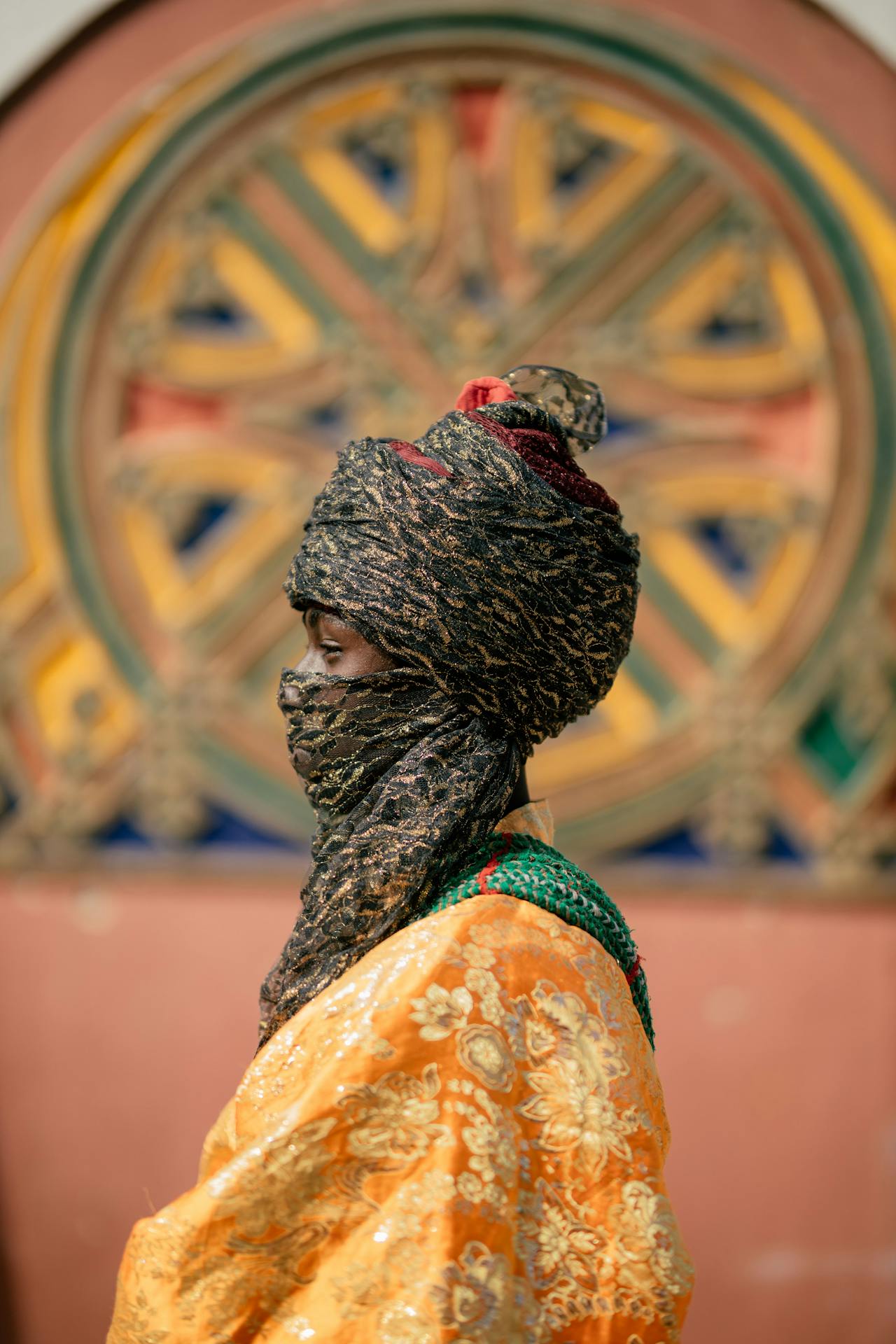 A woman in profile in a gold headwrap and veil and an orange patterned wrap, standing before a painted wheel design on a wall